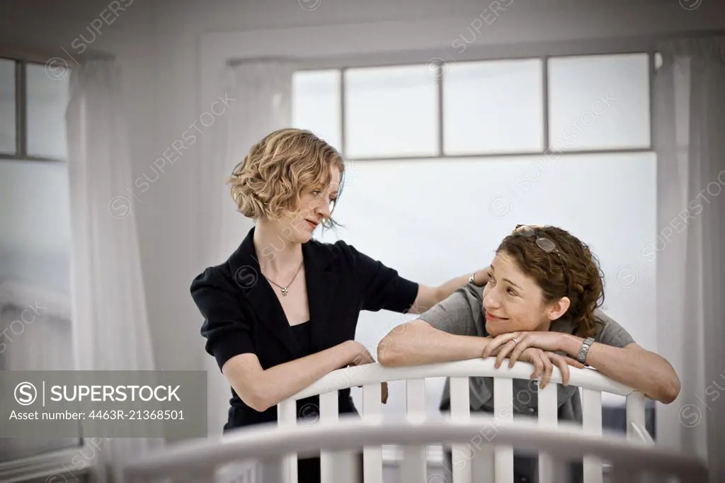 Lesbian couple looking reassuredly at each other while standing beside a cot in a nursery.