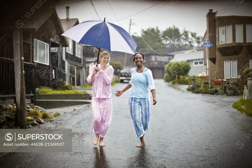 Two laughing young women having fun wearing their pajamas while walking down a residential street in the rain.