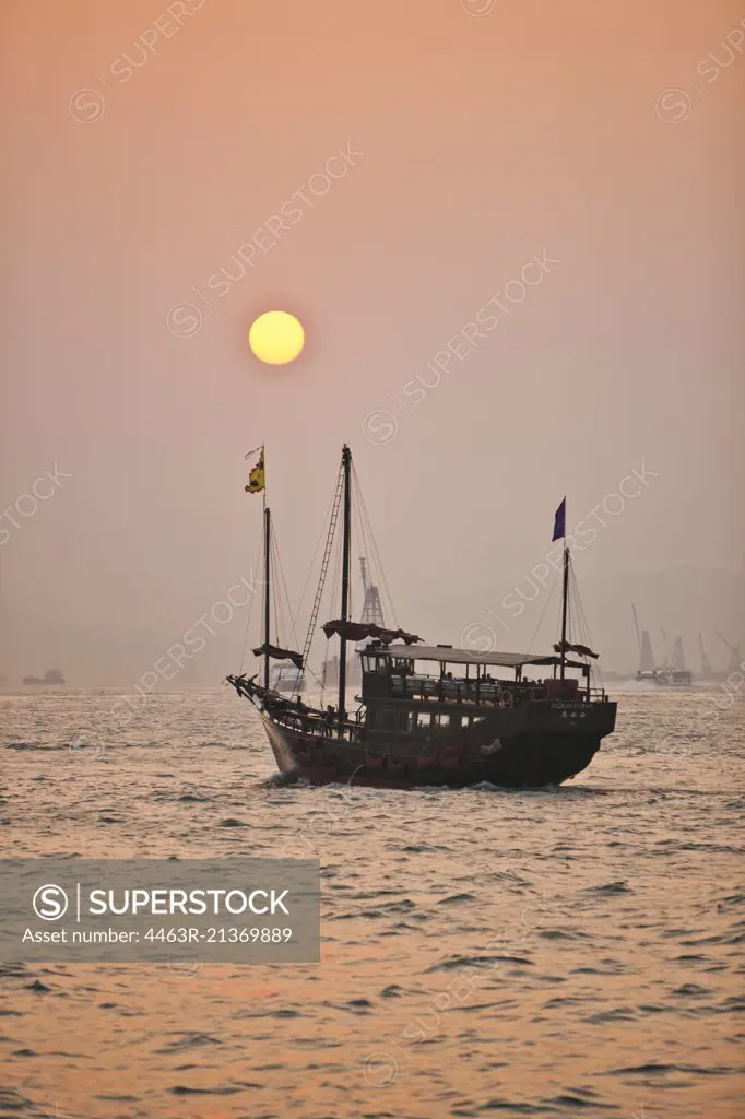 Traditional junk boat sailing on an urban harbor at sunset.