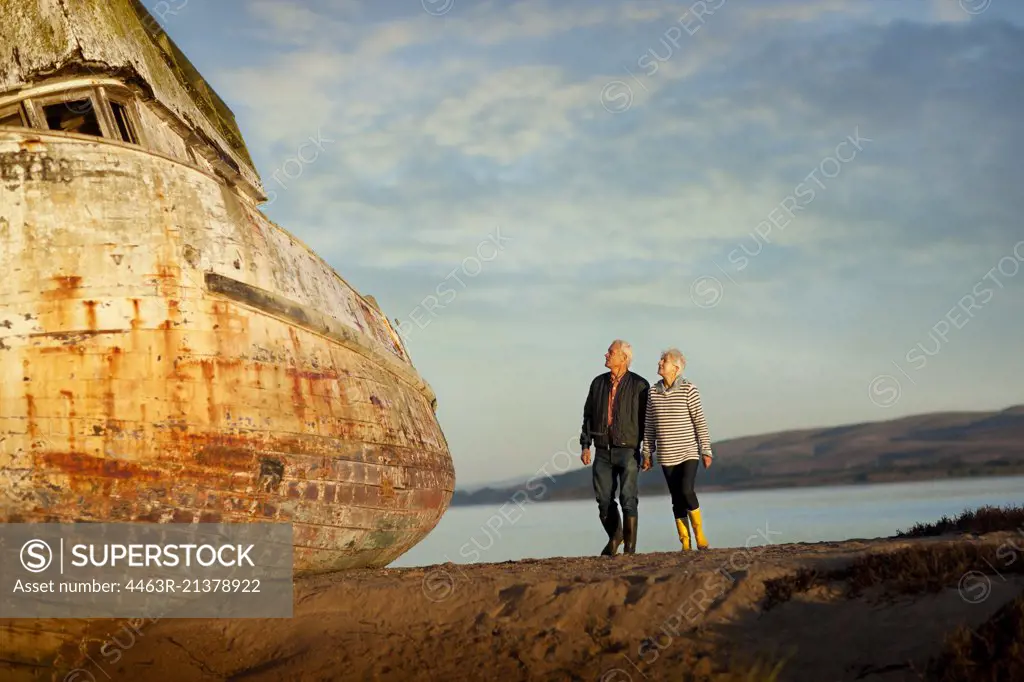 Senior couple walking on the beach hand-in-hand next to an obsolete boat.