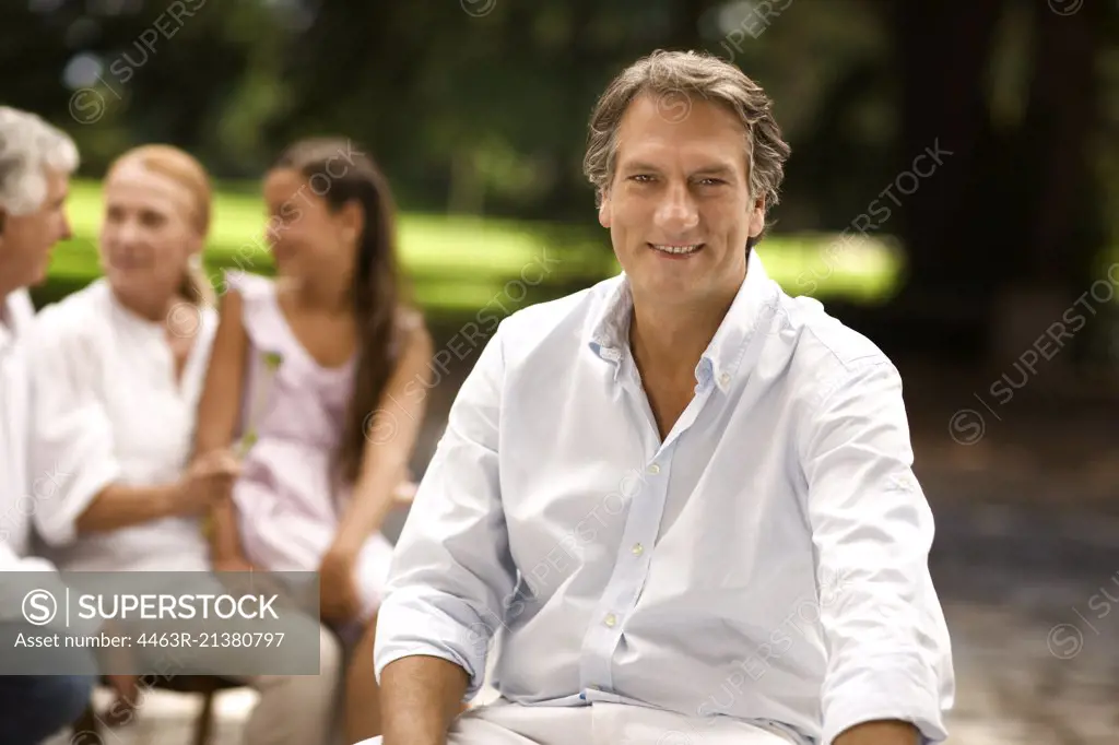 Portrait of mature man sitting outdoors, with family in the background.