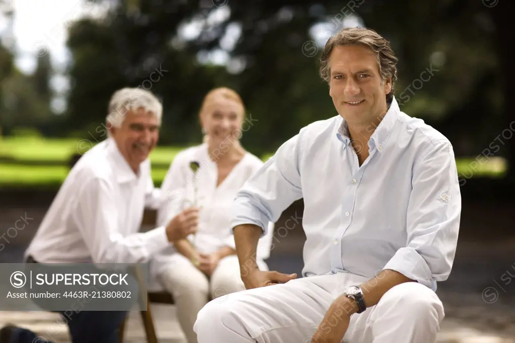 Portrait of mature man sitting outdoors, with family in the background.