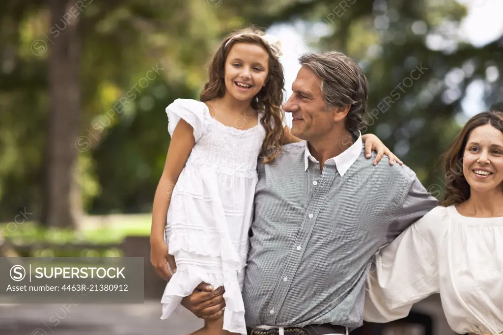 Father holds his daughter in his arms while standing beside his wife.