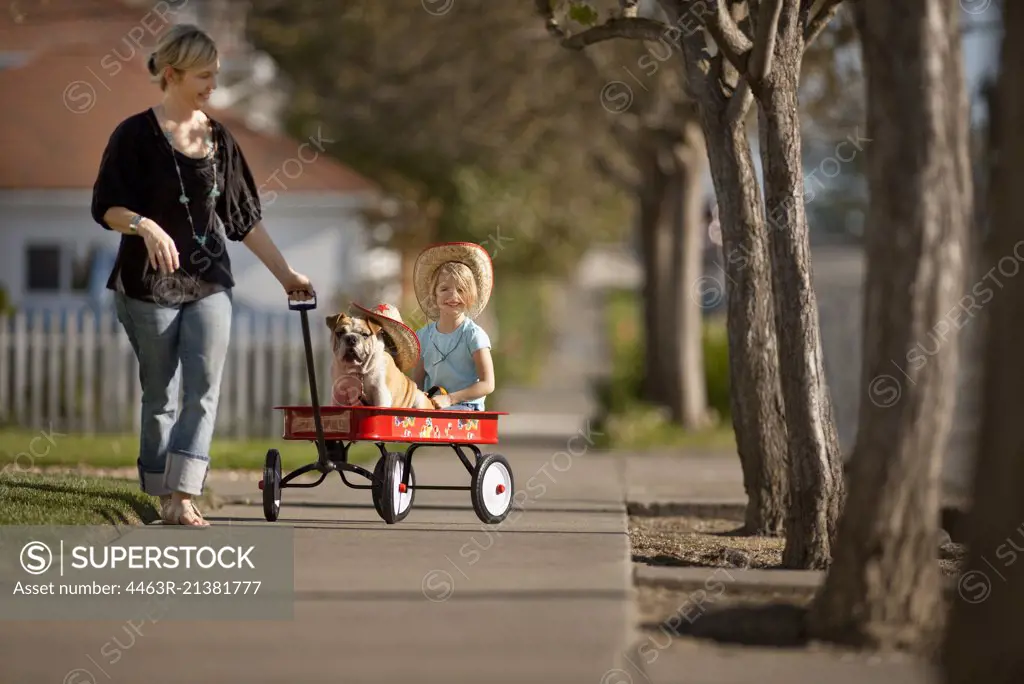 Woman pulling her daughter and a bulldog in a red wagon.