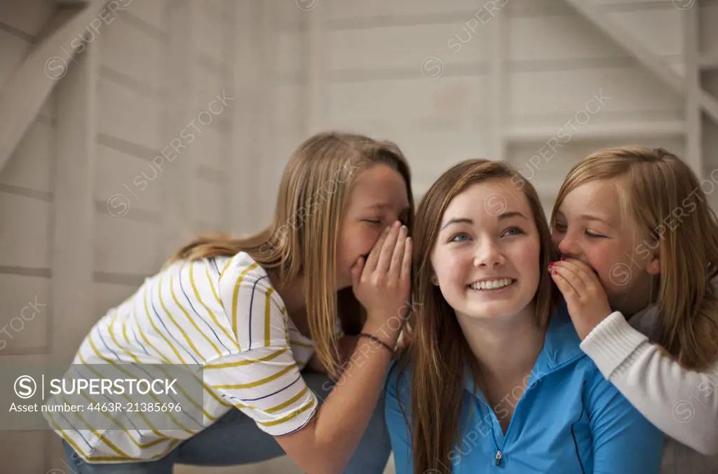 Grinning teenage girl listening while her two younger sisters whisper into both her ears.