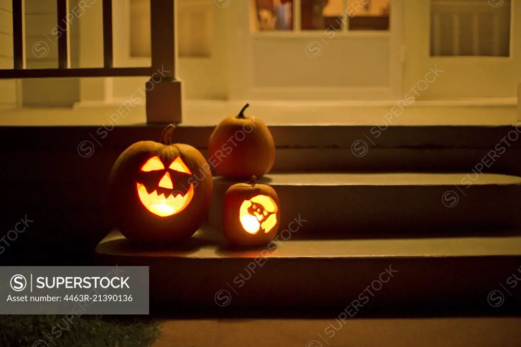 Two jack o' lanterns glowing on the steps of a house beside a normal pumpkin.