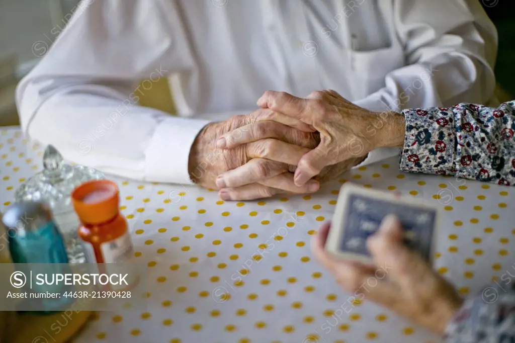 Elderly wife gently places her hand atop her husband's hands as they play cards together at the kitchen table.