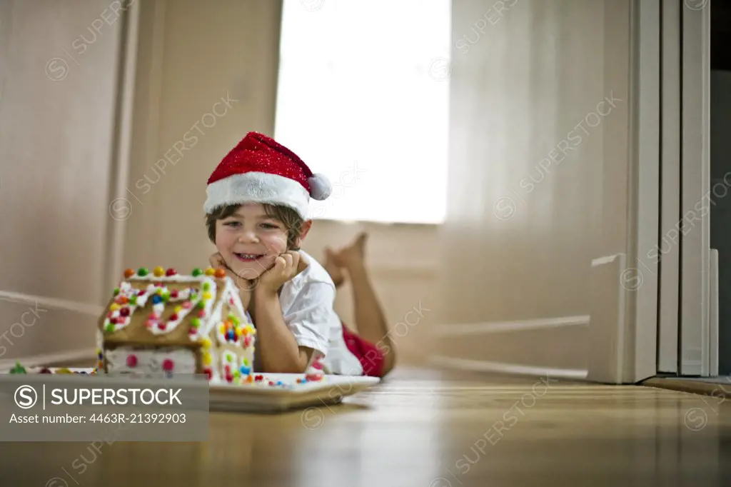 Young boy wearing a Santa hat next to a gingerbread house.