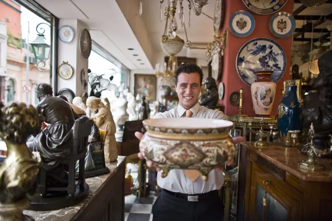 Portrait of a smiling mid-adult businessman holding an antique bowl while standing inside an antique store.