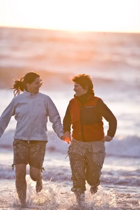 Two women holding hands while running on a beach.