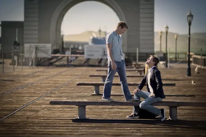 Mid-adult man sitting on a bench looking up at another mid-adult man outside.