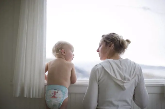 Toddler looking at his young adult mother as they look through a window.