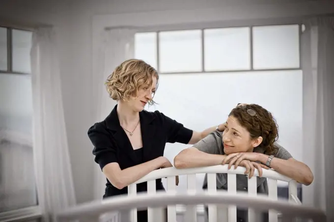 Lesbian couple looking reassuredly at each other while standing beside a cot in a nursery.