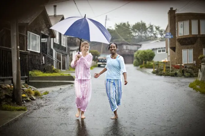 Two laughing young women having fun wearing their pajamas while walking down a residential street in the rain.