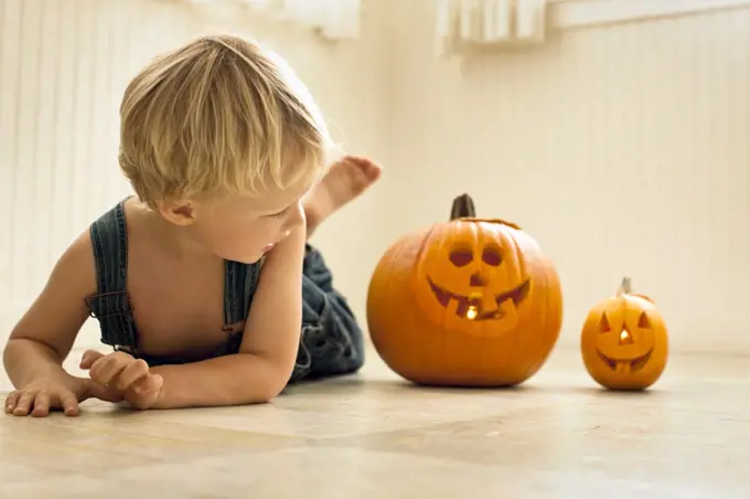 Young boy lies front down on a floor and looks back at a big Jack O'Lantern and a small Jack O'Lantern lit with candles inside next to him as he poses for a portrait.