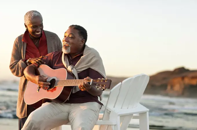 Happy senior woman listening to her husband play an acoustic guitar on a beach.