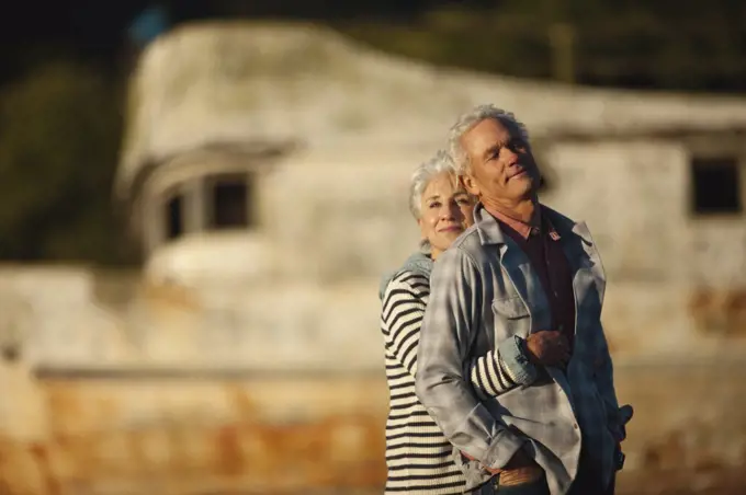 Senior couple with their arms around each other stand on a beach in front of an abandoned boat. 