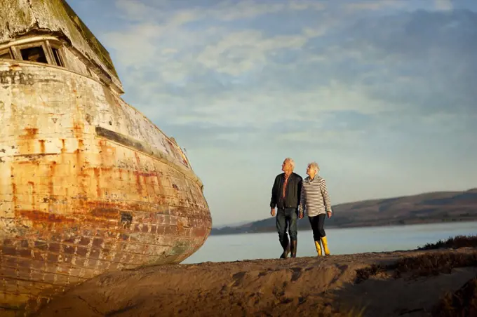 Senior couple walking on the beach hand-in-hand next to an obsolete boat.