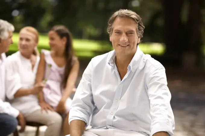 Portrait of mature man sitting outdoors, with family in the background.