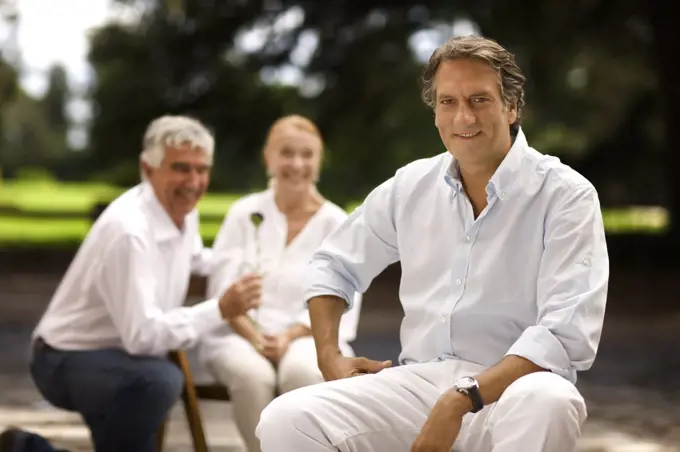 Portrait of mature man sitting outdoors, with family in the background.