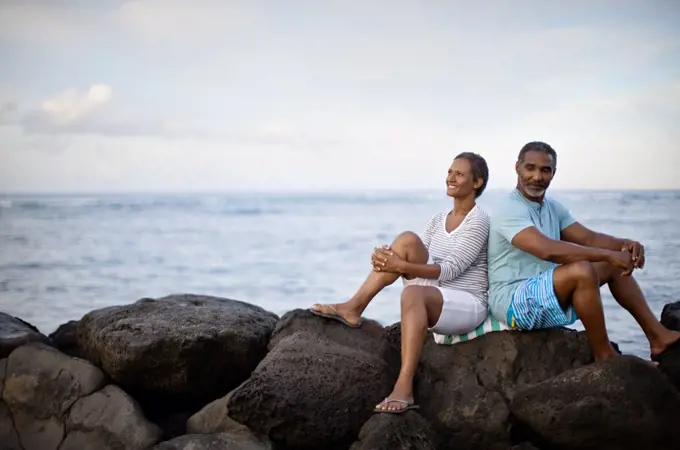 Portrait of mature couple sitting back to back waterside.