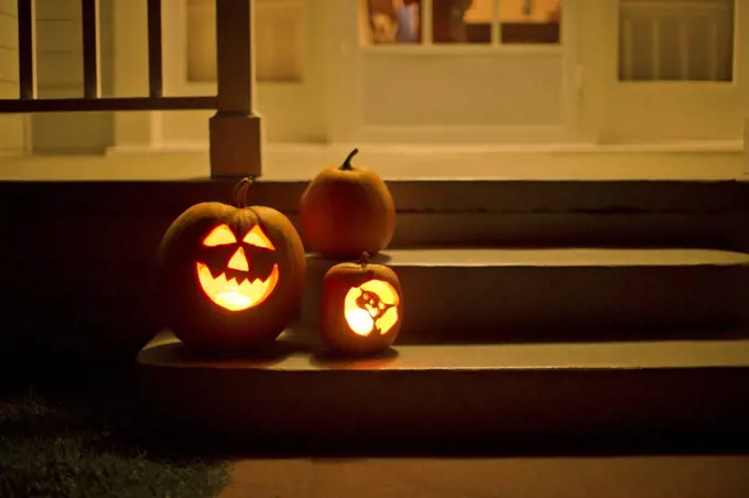 Two jack o' lanterns glowing on the steps of a house beside a normal pumpkin.