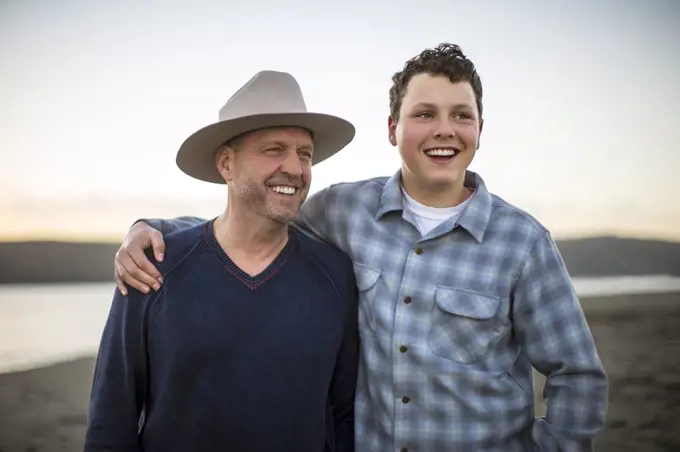 Teenage boy and his dad laughing together on the beach at sunset.