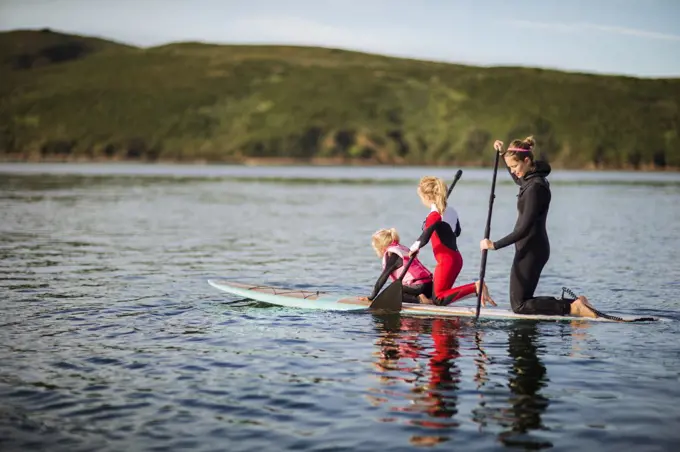 Young woman and her two daughters paddleboarding together on a lake.