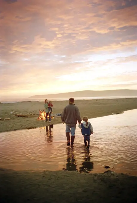 Father holding hands and walking with his young daughter towards a bonfire beside a tidal pool on a sandy beach at sunset.