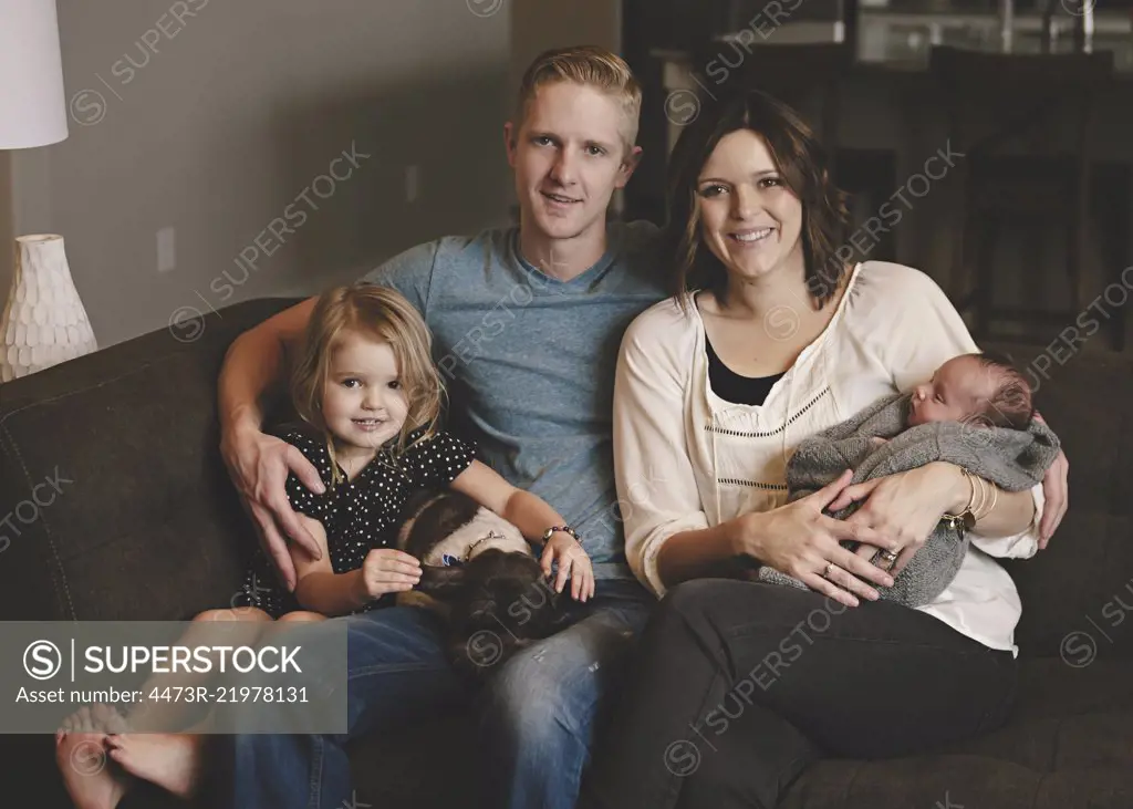 Family group, a couple, mother holding a new baby in her arms, father, daughter and a dog sitting on a sofa.