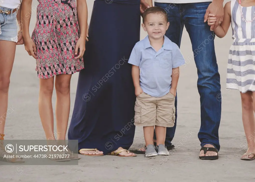 A family group on the beach, waist down, and a small boy in the foreground looking at the camera. 