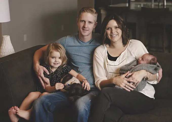 Family group, a couple, mother holding a new baby in her arms, father, daughter and a dog sitting on a sofa.