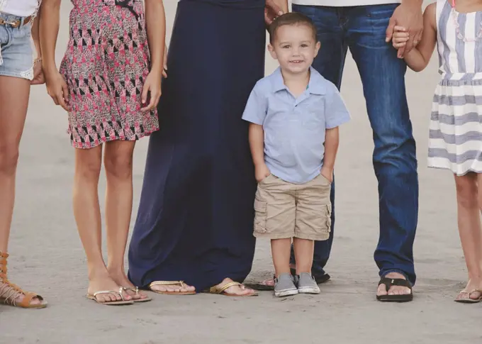 A family group on the beach, waist down, and a small boy in the foreground looking at the camera. 