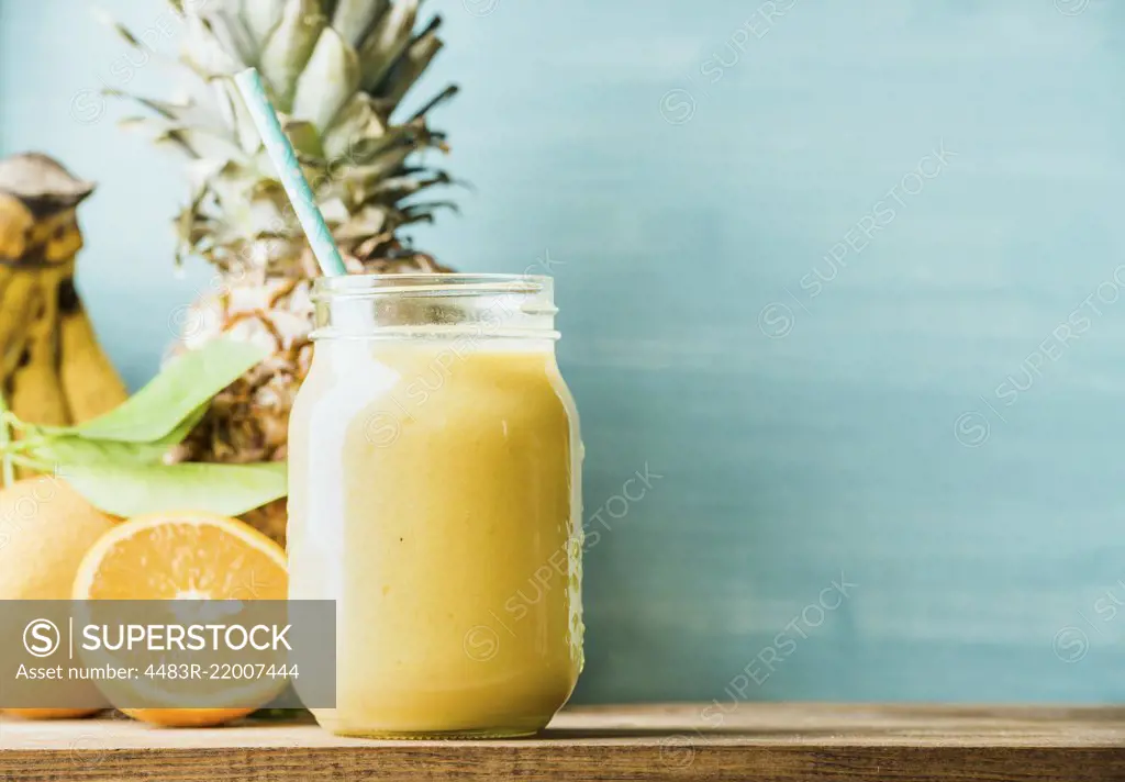 Freshly blended yellow and orange fruit smoothie in glass jar with straw. Turquoise blue background, copy space