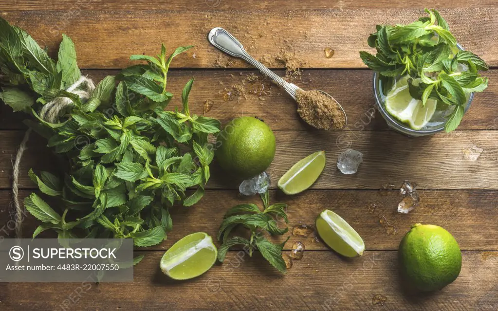 Ingredients for making mojito summer cocktail. Fresh mint bunch, limes, brown sugar and ice over rustic wooden background, top view