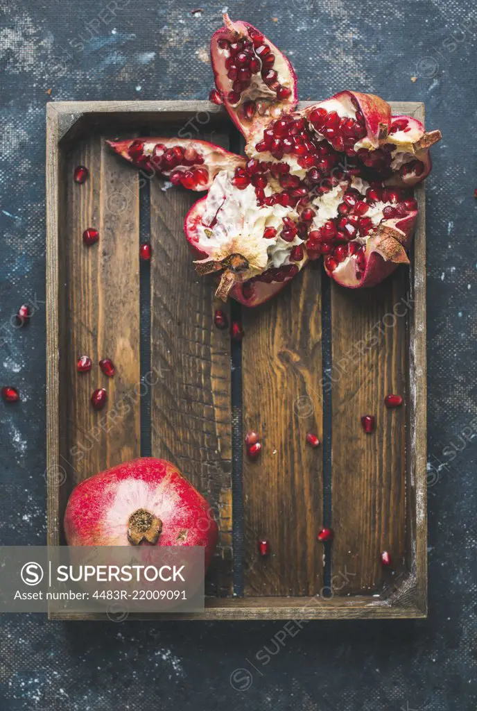 Fresh ripe pomegranates cut in pieces in rustic wooden tray over dark blue plywood background, top view, copy space, vertical composition