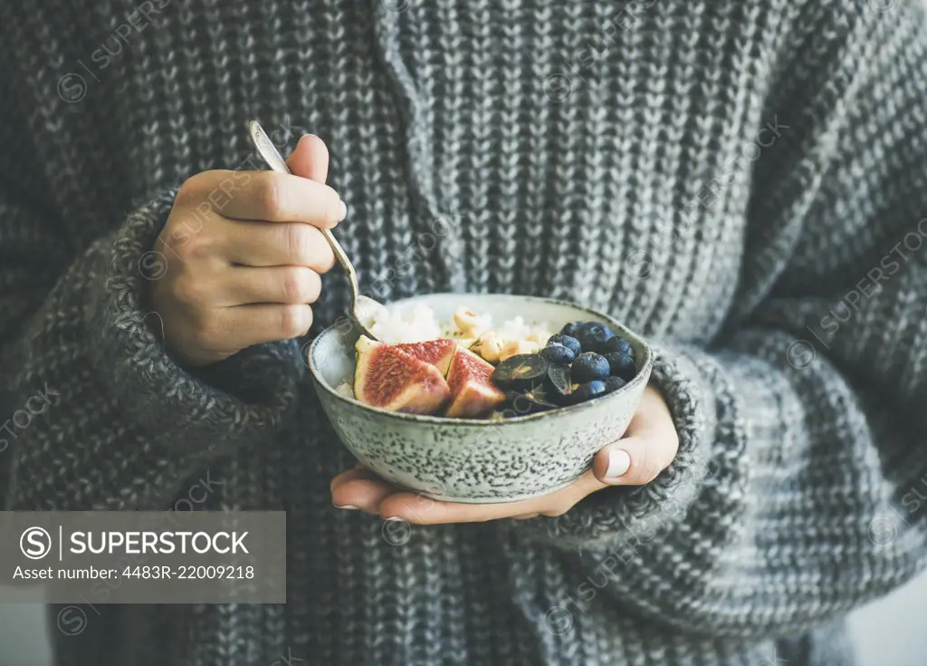 Healthy winter breakfast. Woman in grey woolen sweater eating rice coconut porridge with figs, berries and hazelnuts. Clean eating, vegetarian, vegan, alkiline diet food concept