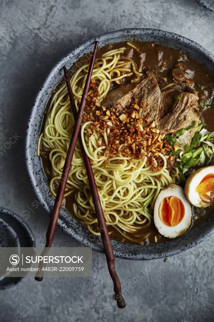 Vertical close up shot of japanese ramen with pork belly, mushrooms and marinated eggs served in blue handmade ceramic bowl garnished with chives