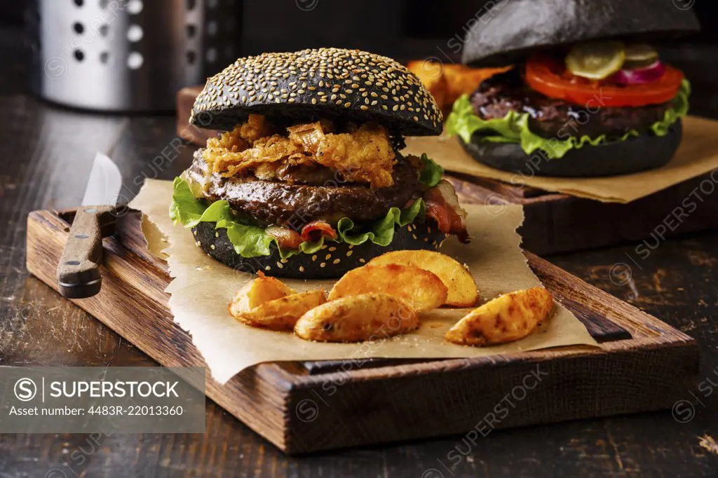 Black burger with sesame seed bun, meat, bacon, onion fries rings and potato wedges on dark wooden background