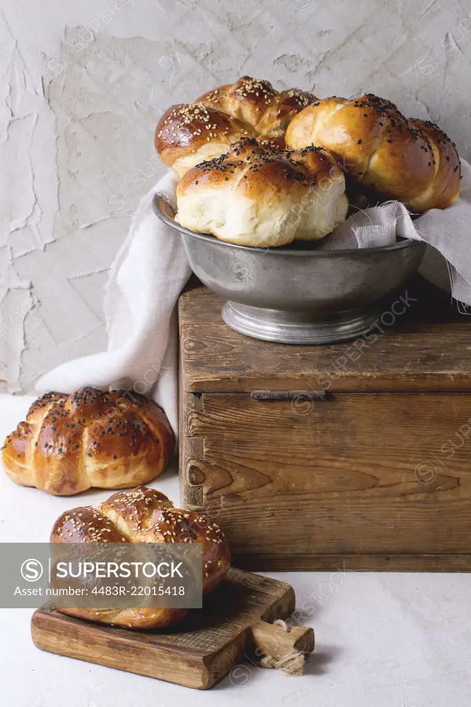 Heap of sweet round sabbath challah bread with white and black sesame seeds in vintage metal bowl on wooden chest and on small cutting board over white table with plastered wall at background.