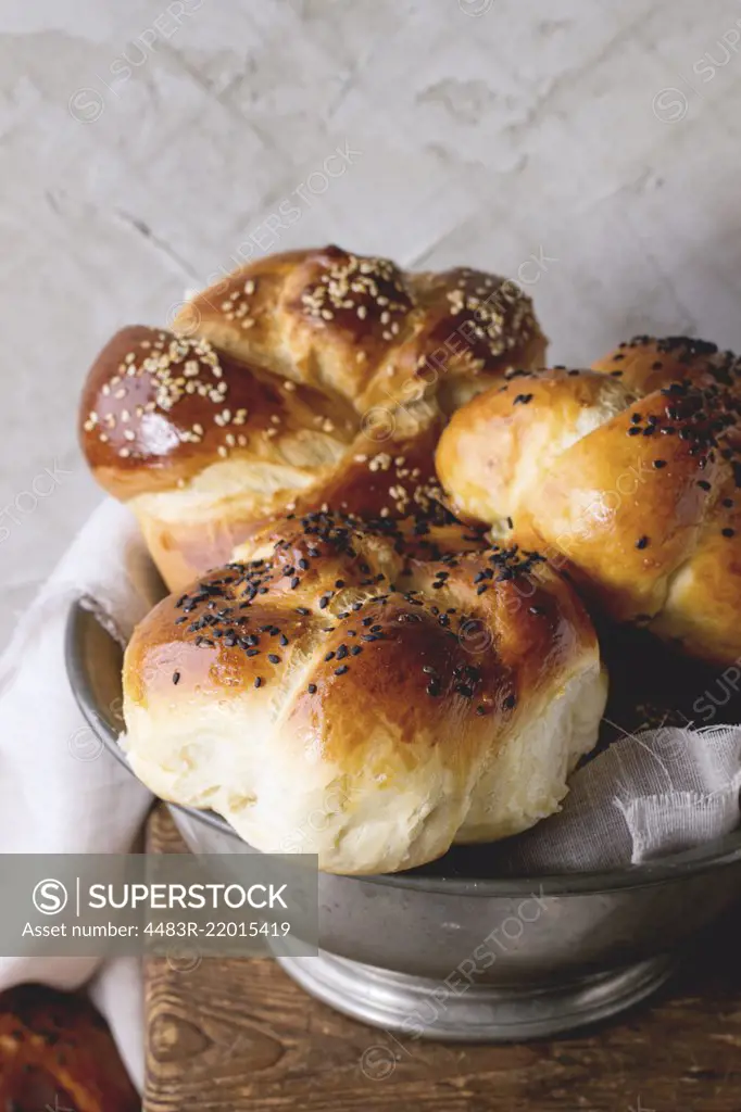 Heap of sweet round sabbath challah bread with white and black sesame seeds in vintage metal bowl on wooden table with plastered wall at background.