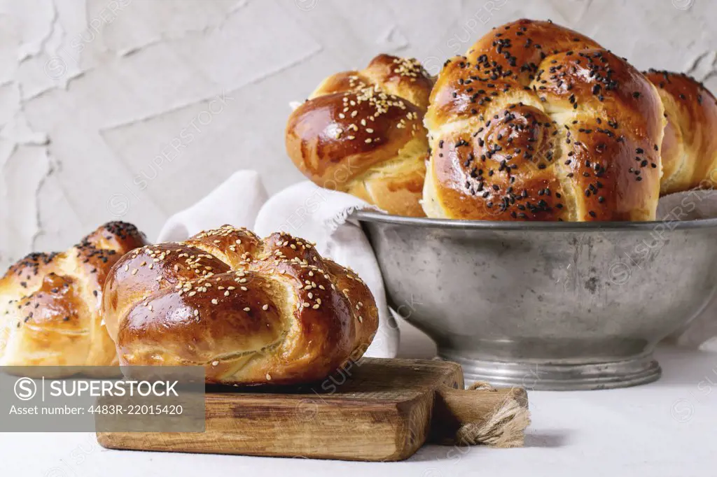 Heap of sweet round sabbath challah bread with white and black sesame seeds in vintage metal bowl and on small cutting board over white table with plastered wall at background.