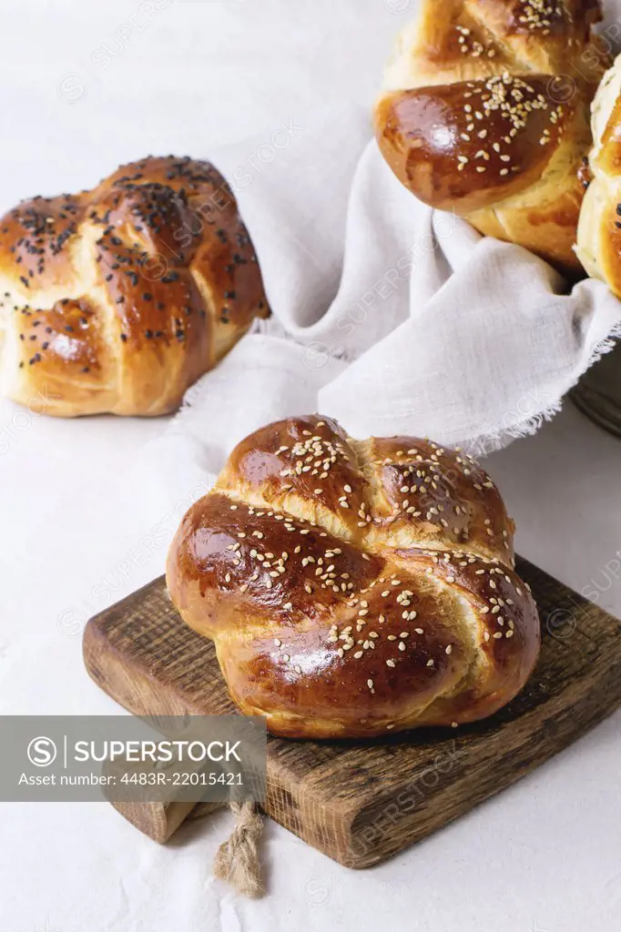 Heap of sweet round sabbath challah bread with white and black sesame seeds on small cutting board over white table with plastered wall at background.