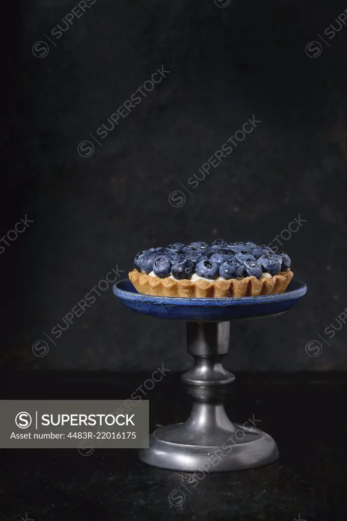 Lemon tartlet with fresh blueberries, served on vintage cake stand over black background.