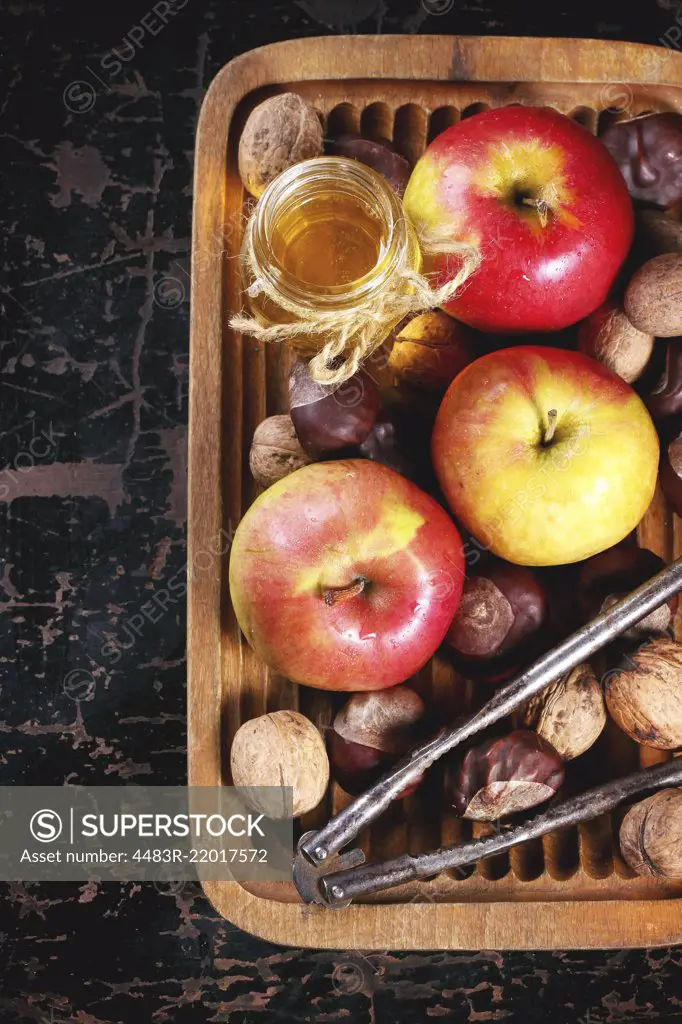 Glass jar of honey with apples, chestnuts and walnuts in wooden plate over black wooden table. Top view