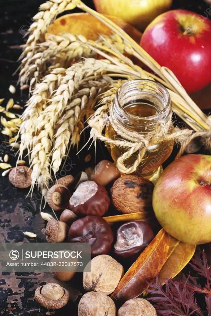 Glass jar of honey with apples, ears of wheat, chestnuts and walnuts over black wooden table