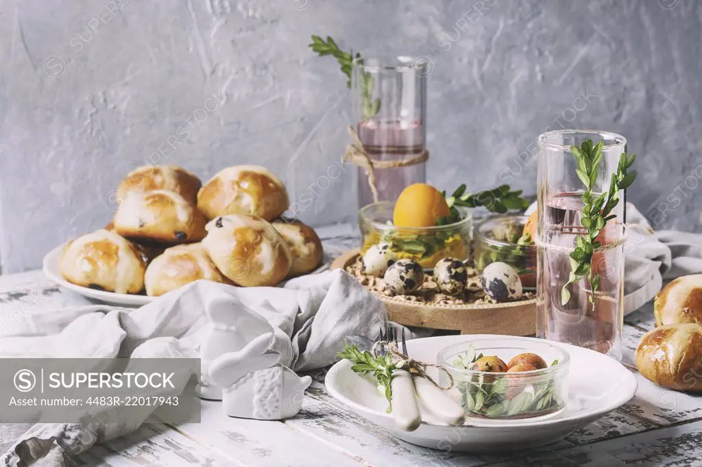 Easter table setting with colored orange eggs, hot cross buns, green branches decorated, empty white plate with cutlery, glass of lemonade drink over white plank wooden table with textile tablecloth.