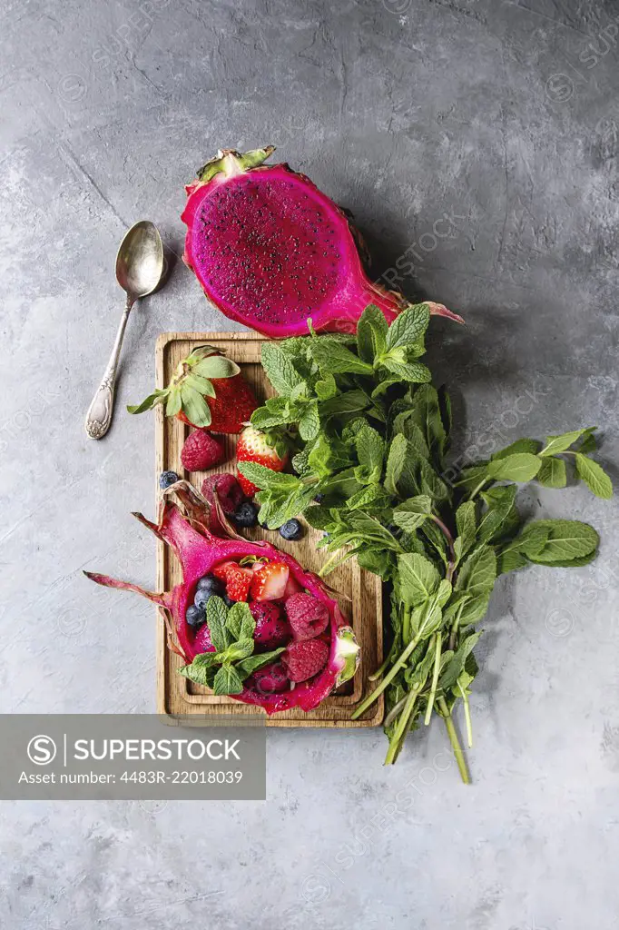 Vegan fruit salad with berries and mint served in pink dragon fruit with ingredients above on wooden serving board over blue texture background. Top view, space.