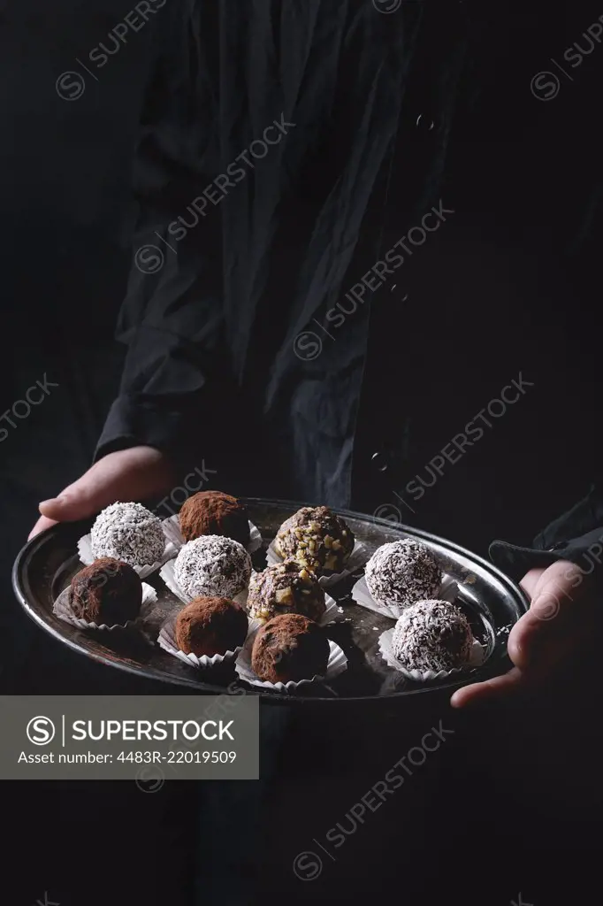 Variety of homemade dark chocolate truffles with cocoa powder, coconut, walnuts on vintage tray in kid's hands in black shirt. Dark background.