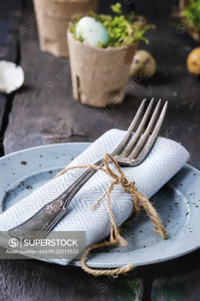 Table setting decor colorful Easter quail eggs with spring cherry flowers, moss in garden pots, empty plates, cutlery over old wood background. Dark rustic style.